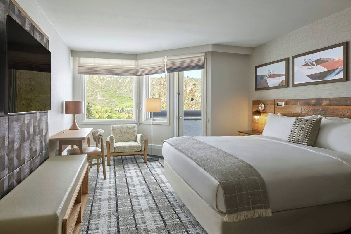 Une chambre d’hôtel avec un grand lit, un bureau avec une chaise, une petite table et une fenêtre donnant sur un paysage verdoyant.