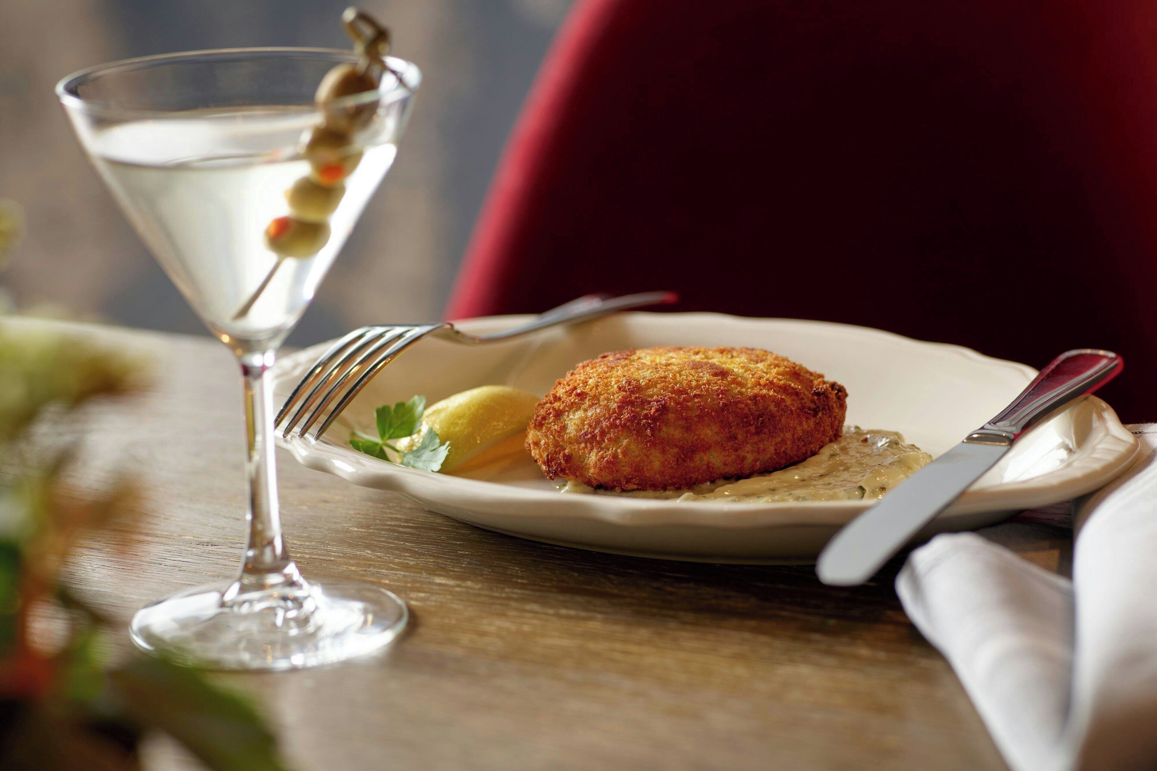Ein Martini-Glas mit Cocktailolive, ein Teller mit einer panierten Speise und einem Zitronenschnitzchen sowie Besteck auf einer Holztischplatte.