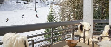 A balcony with wicker furniture, a round table, and a view of a ski slope with skiers.