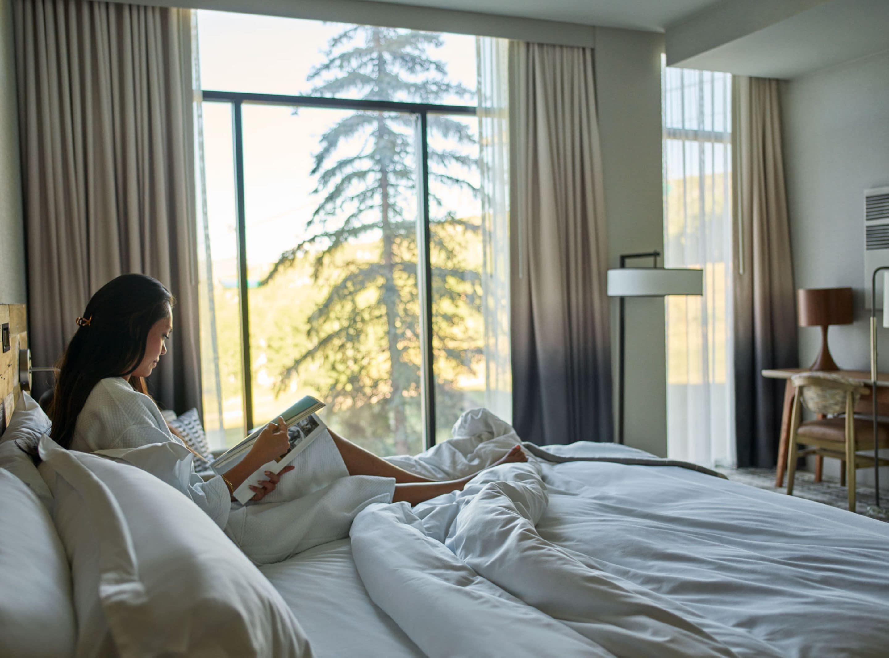 A hotel room with a large window, a bed, a bedside table, and a lamp.