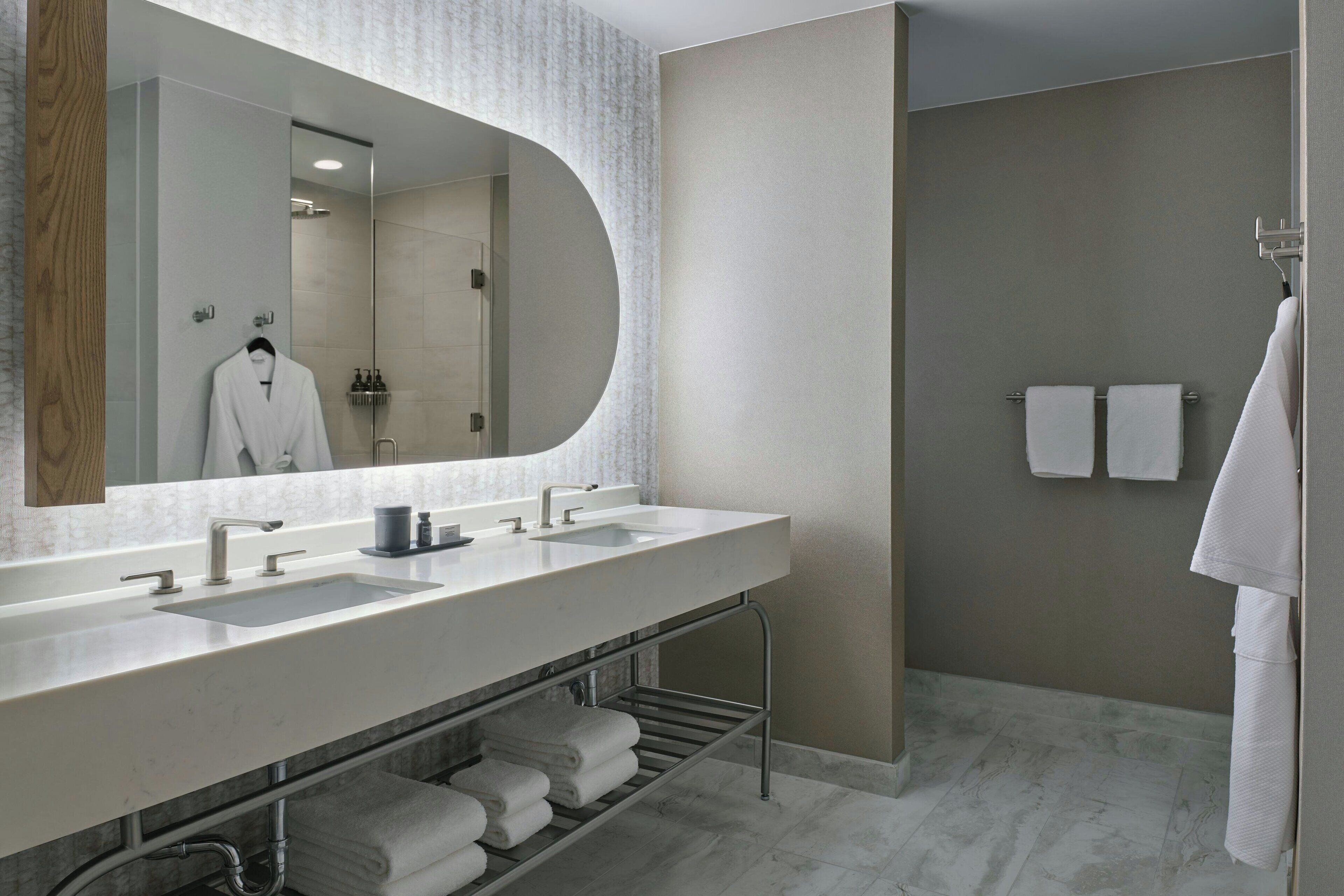 A modern bathroom with a large mirror, two sinks, a towel rack, and a shower area.