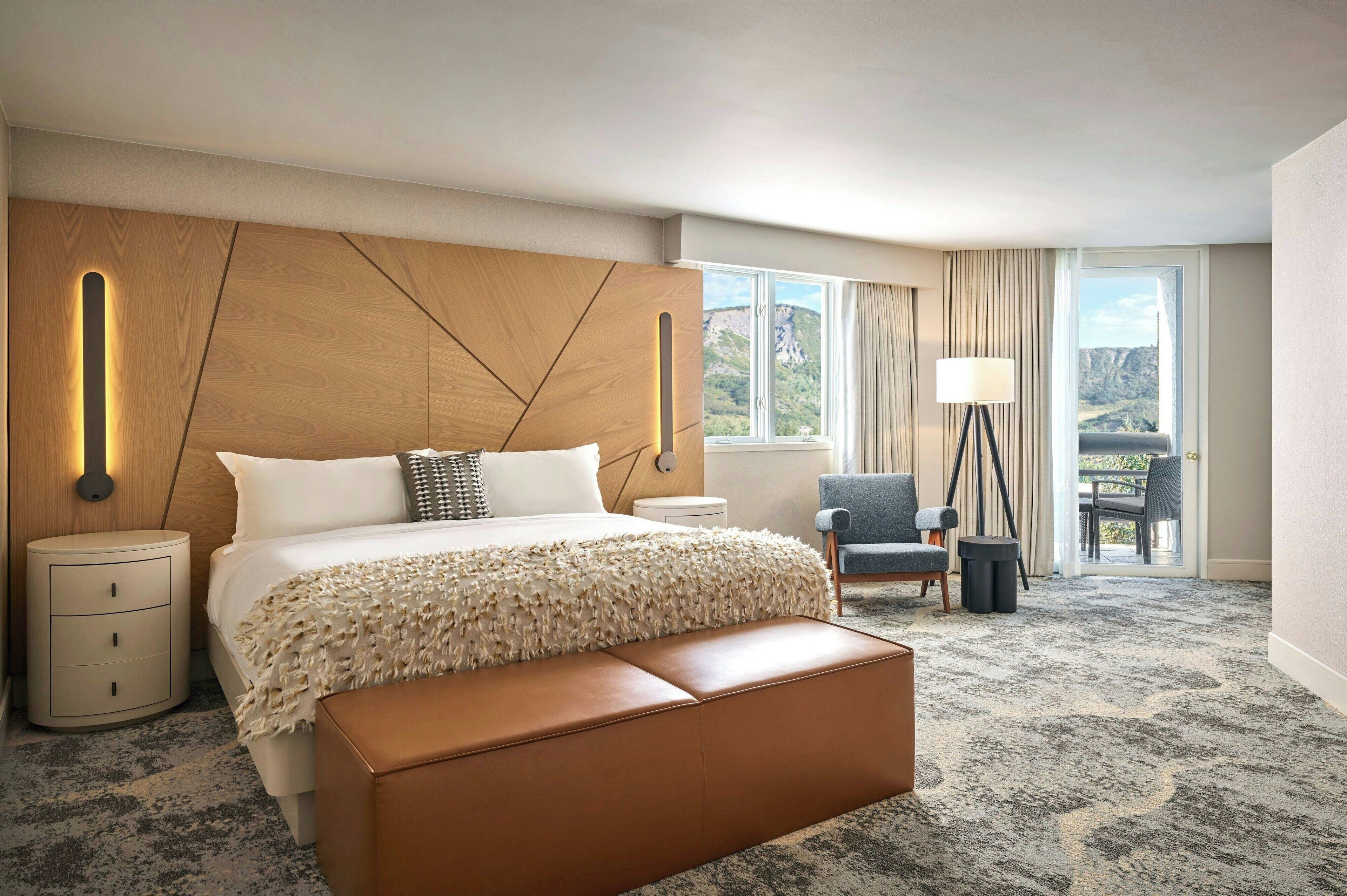 A hotel room with a large bed, a wooden headboard, a view of the outdoors, a chair, and a lamp.