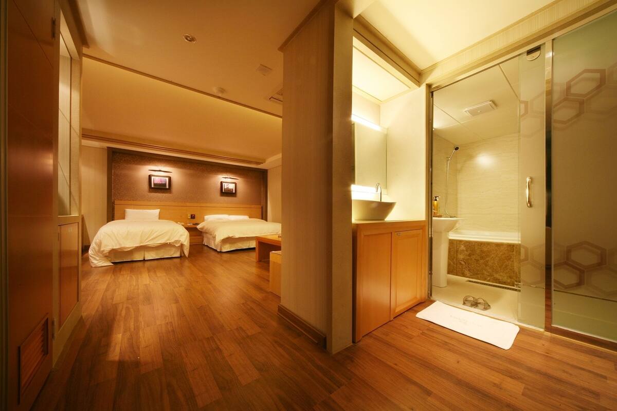 A hotel room with two beds, a glass shower enclosure, and wooden flooring.