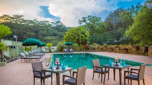 A poolside area with lounge chairs, a dining table set for two, and a view of a mountain backdrop. - Hunas Falls Hotel (Ukuwela)