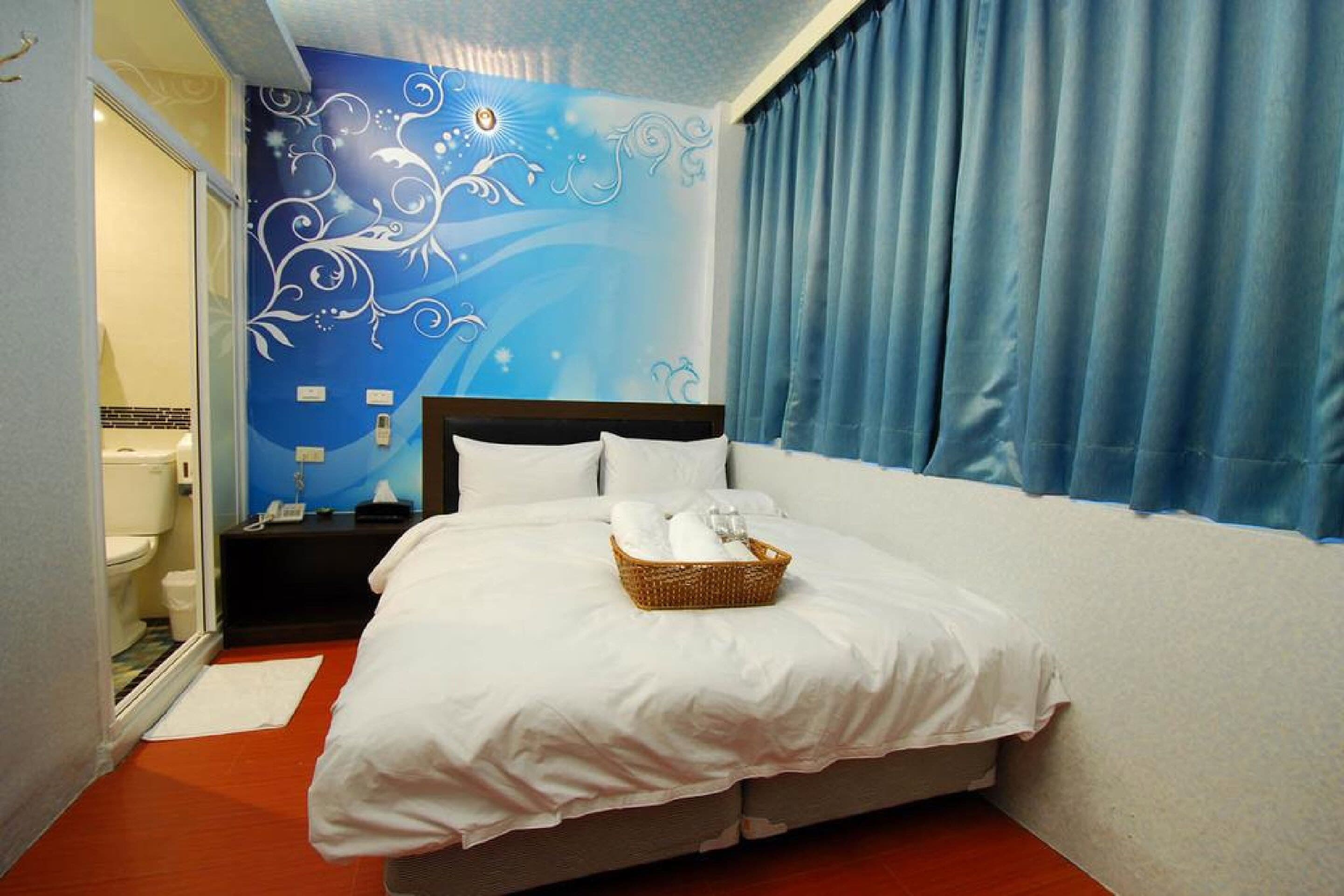 A hotel room with a bed, a nightstand, a wall with a decorative mural, a bathroom visible through an open door, and a blue curtain.