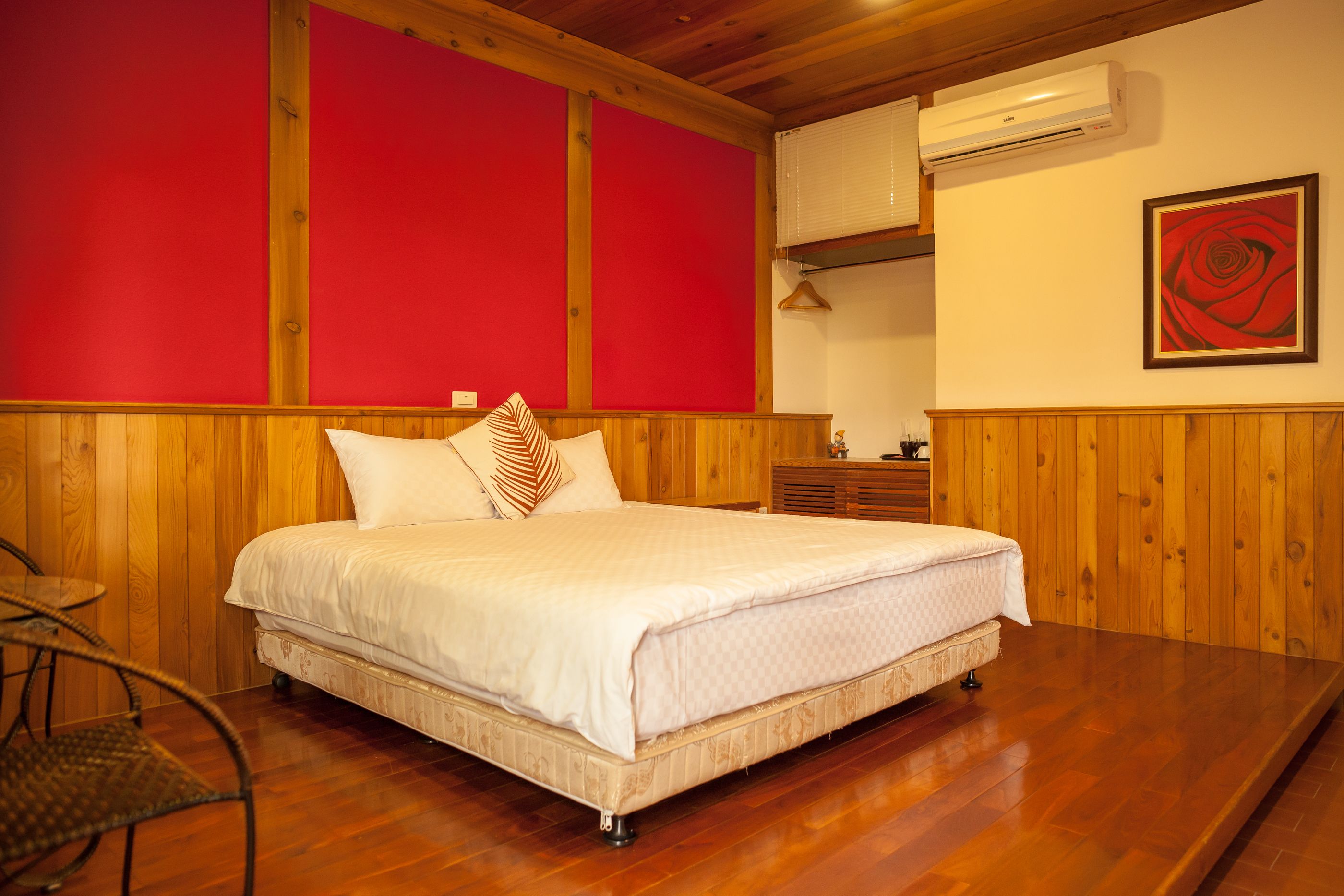 Un dormitorio con cabecera de madera, cama con ropa blanca, pared roja y un cuadro enmarcado de una rosa.