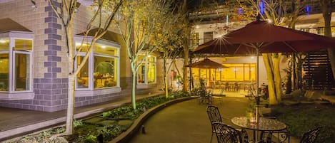 Un patio al aire libre con mesas y sillas, rodeado de árboles y un edificio con ventanas amplias.