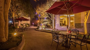 An outdoor patio area with tables and chairs, surrounded by trees and illuminated by ambient lighting.