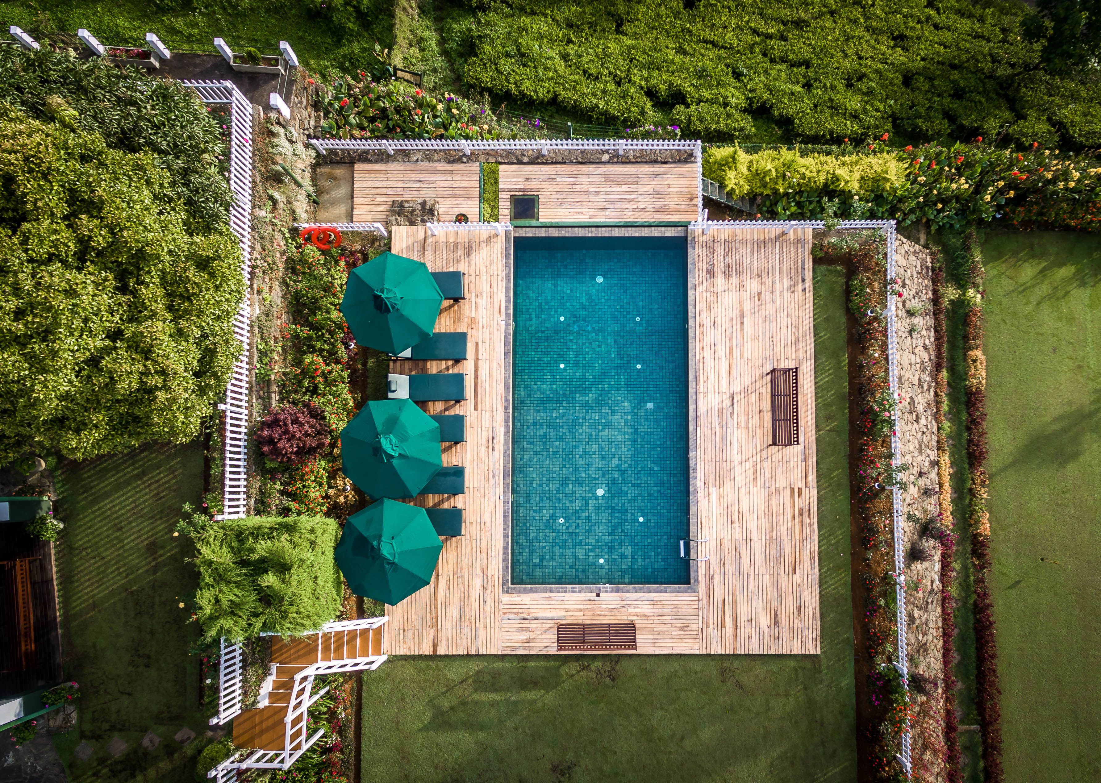 An aerial view of a rectangular swimming pool with a wooden deck, surrounded by lush greenery and a white fence.