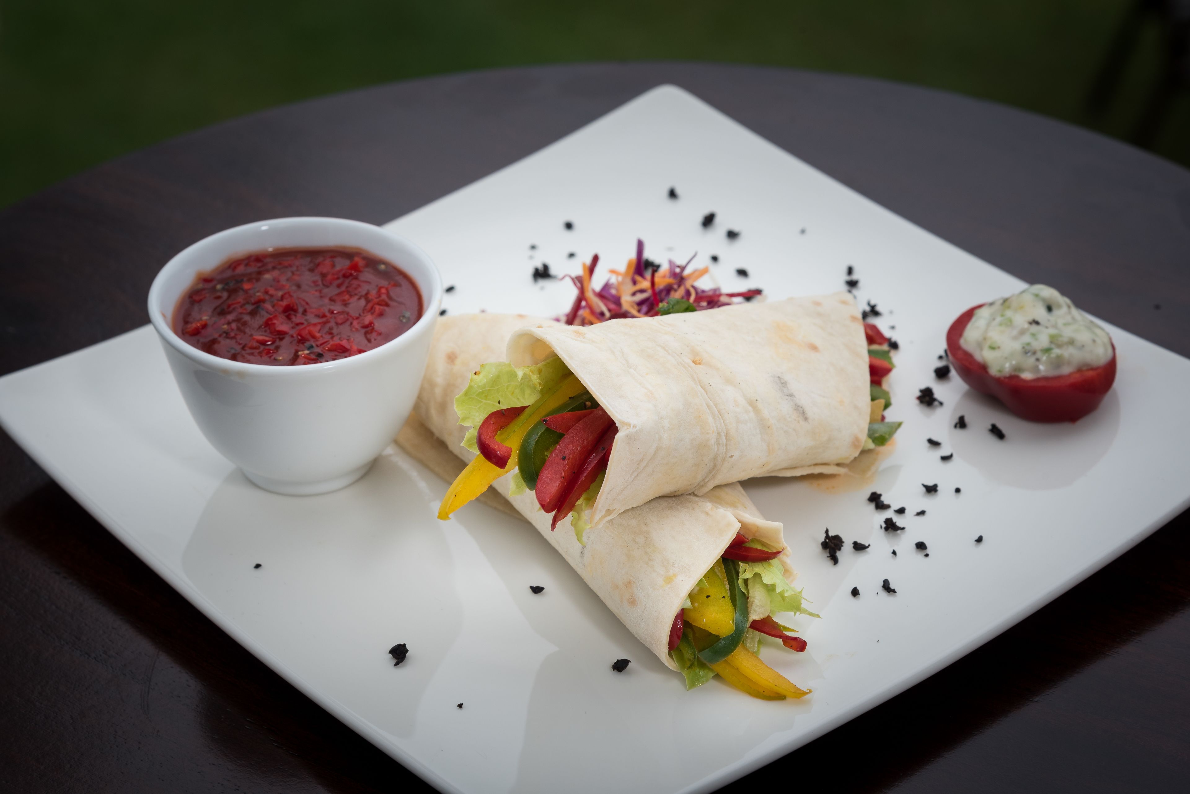 A plate with a wrap, a bowl of sauce, and a stuffed tomato.