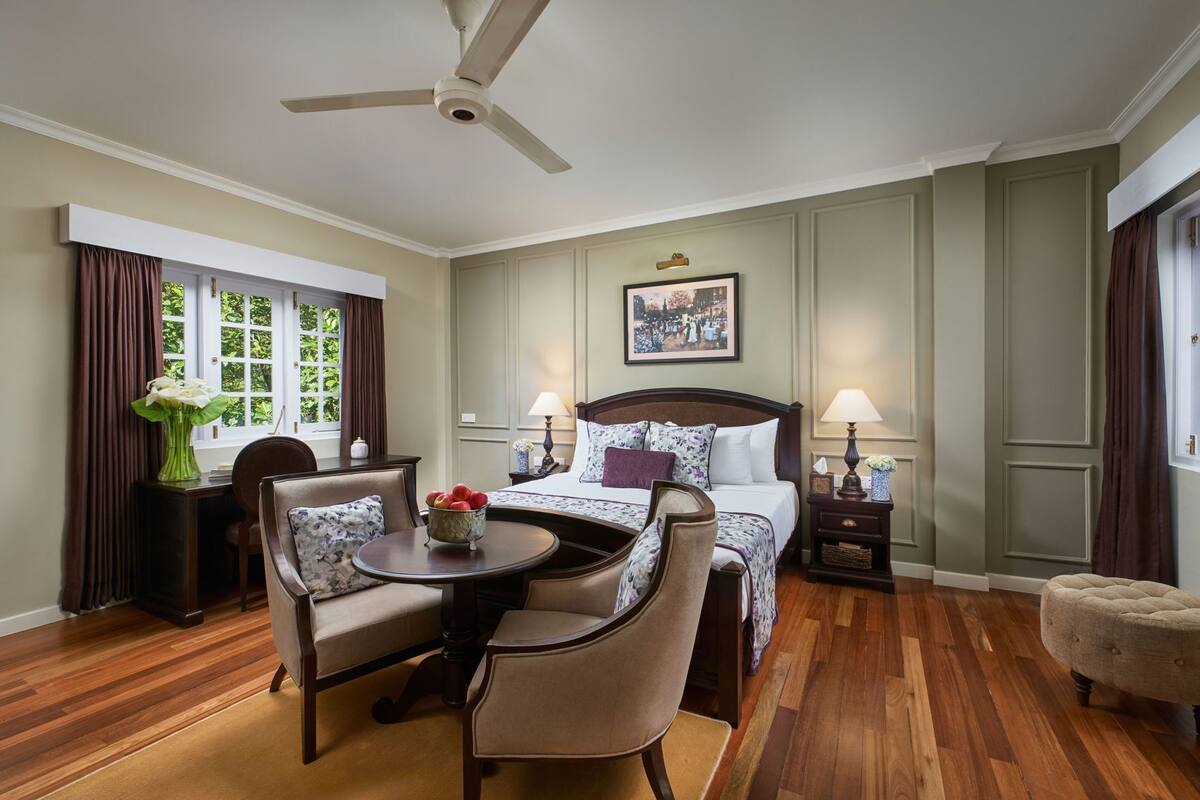 Ein Hotelzimmer mit einem Bett, einem Schreibtisch mit Stuhl, einem runden Tisch mit Stühlen, einem Deckenventilator und einem Fenster mit Vorhängen.