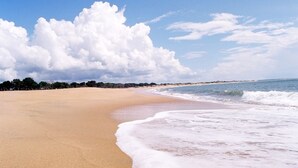 Une vaste plage de sable fin, avec des vagues douces, un ciel dégagé et une ligne d’arbres à l’horizon.