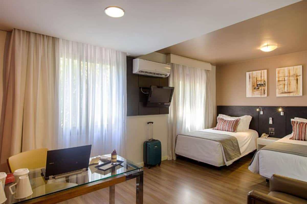 Habitación de hotel con dos camas, un escritorio, una laptop, una maleta y un equipo de aire acondicionado.