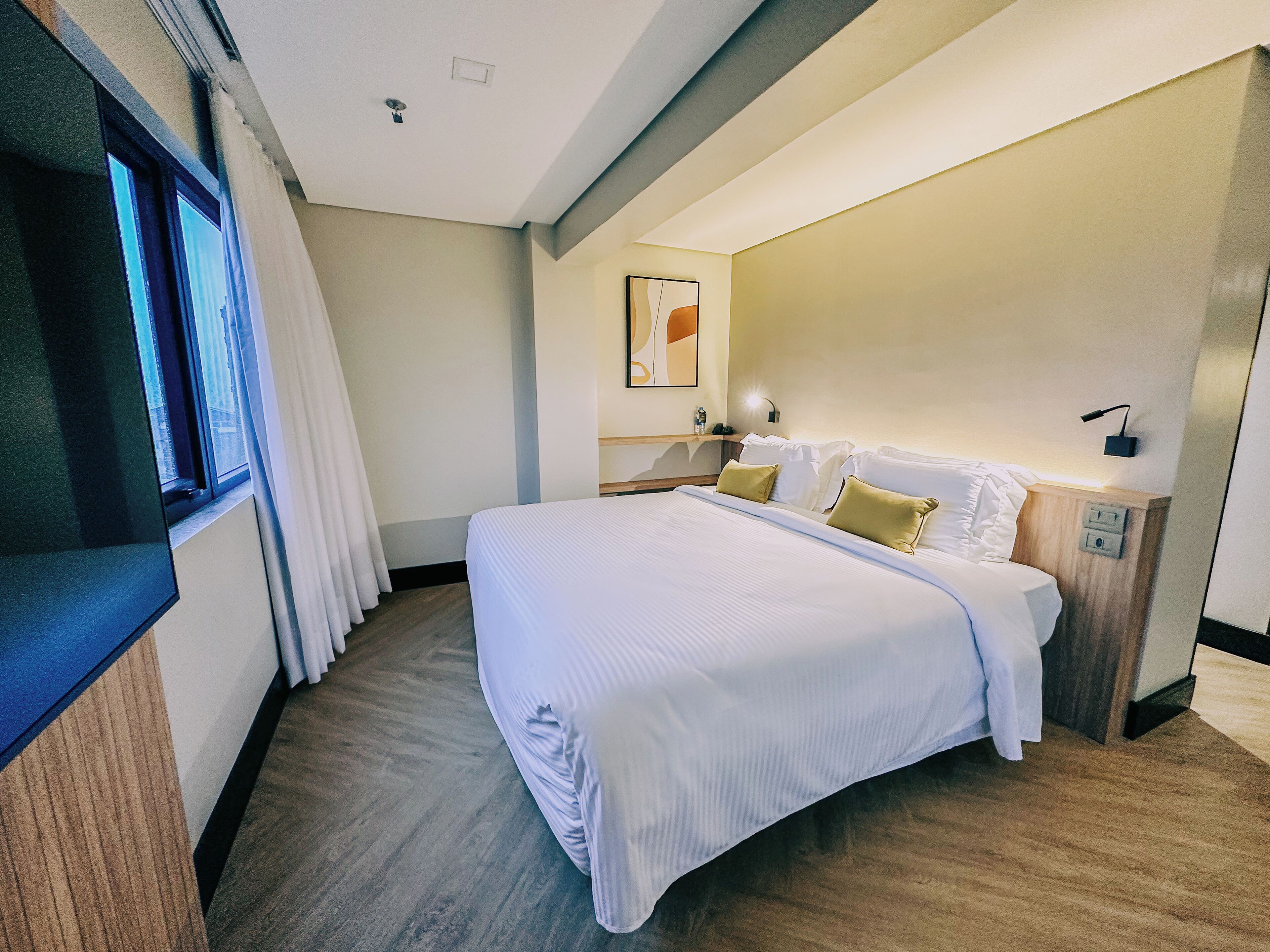 A modern hotel room with a large bed, a flat-screen TV, a bedside table, and a wall-mounted lamp.