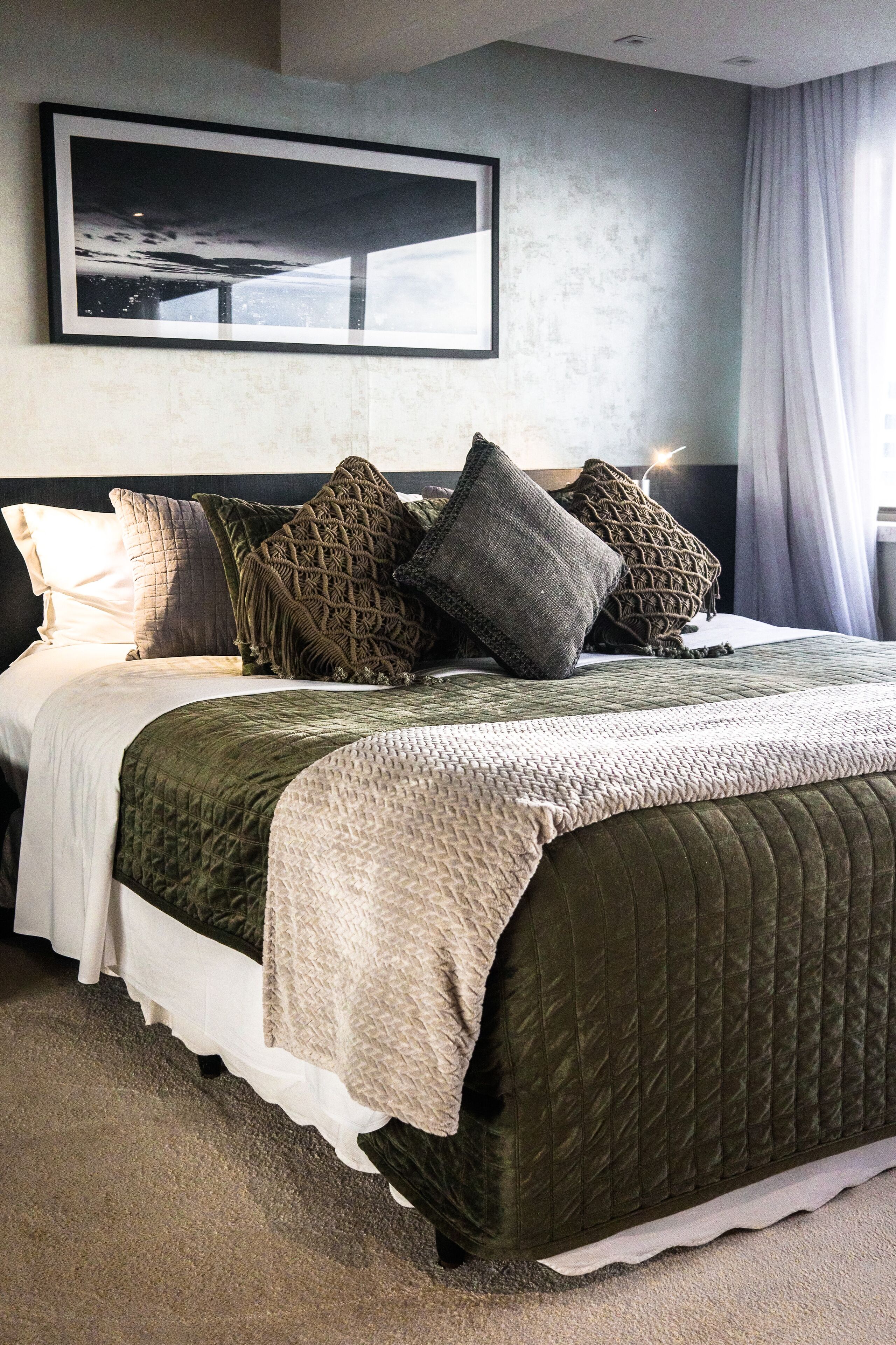 A neatly made bed with a quilted green bedspread and multiple pillows, a framed picture on the wall, and a window with curtains.