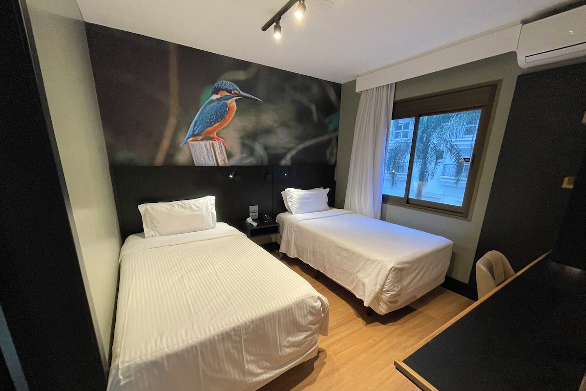 Habitación de hotel con dos camas individuales, un escritorio y un ventanal grande.