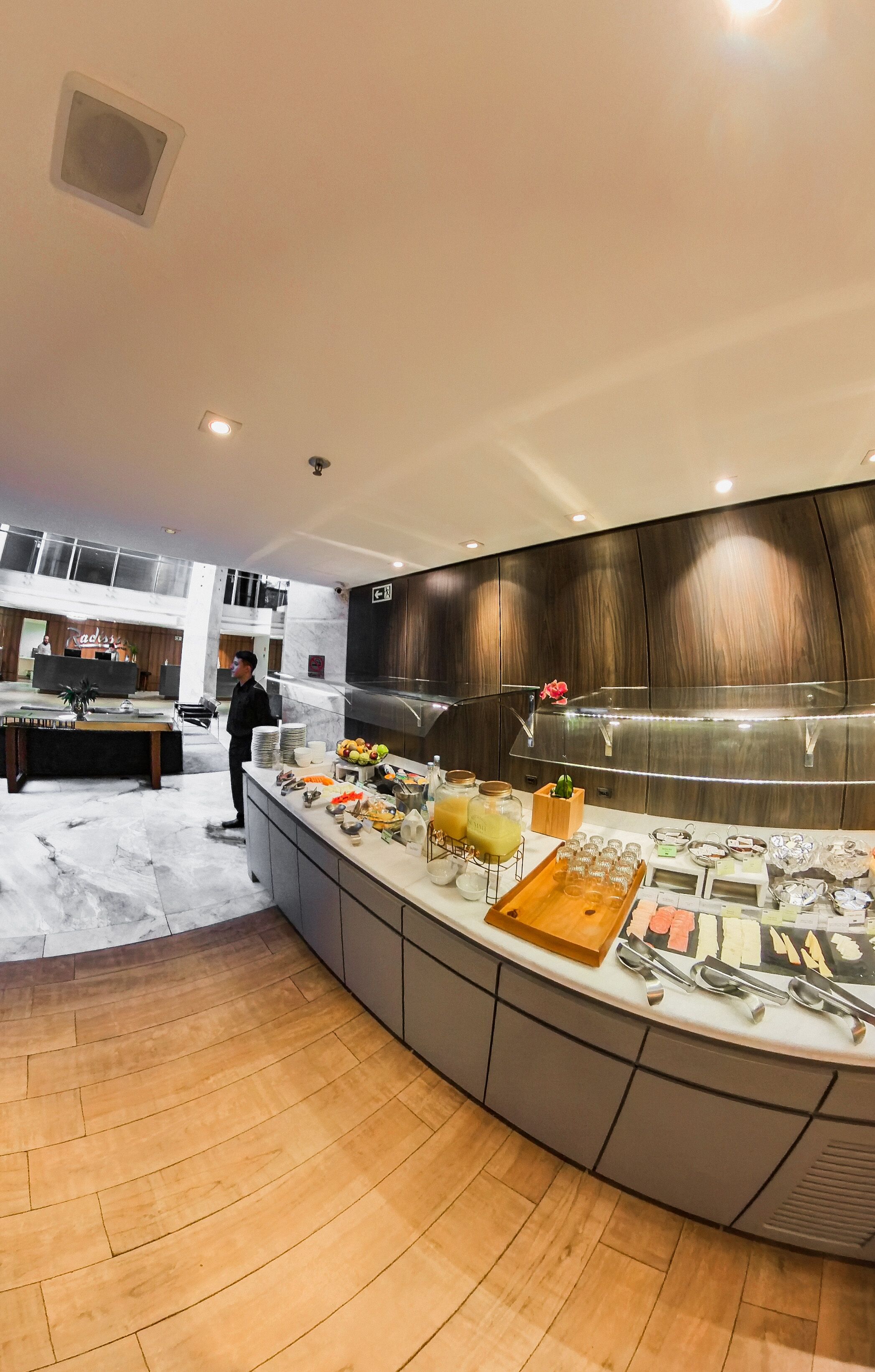 A modern hotel lobby with a curved reception desk, a buffet area with various food items, and a wooden floor.