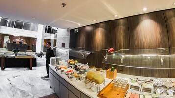 A modern hotel lobby with a curved reception desk, a buffet area with various food items, and a wooden floor.