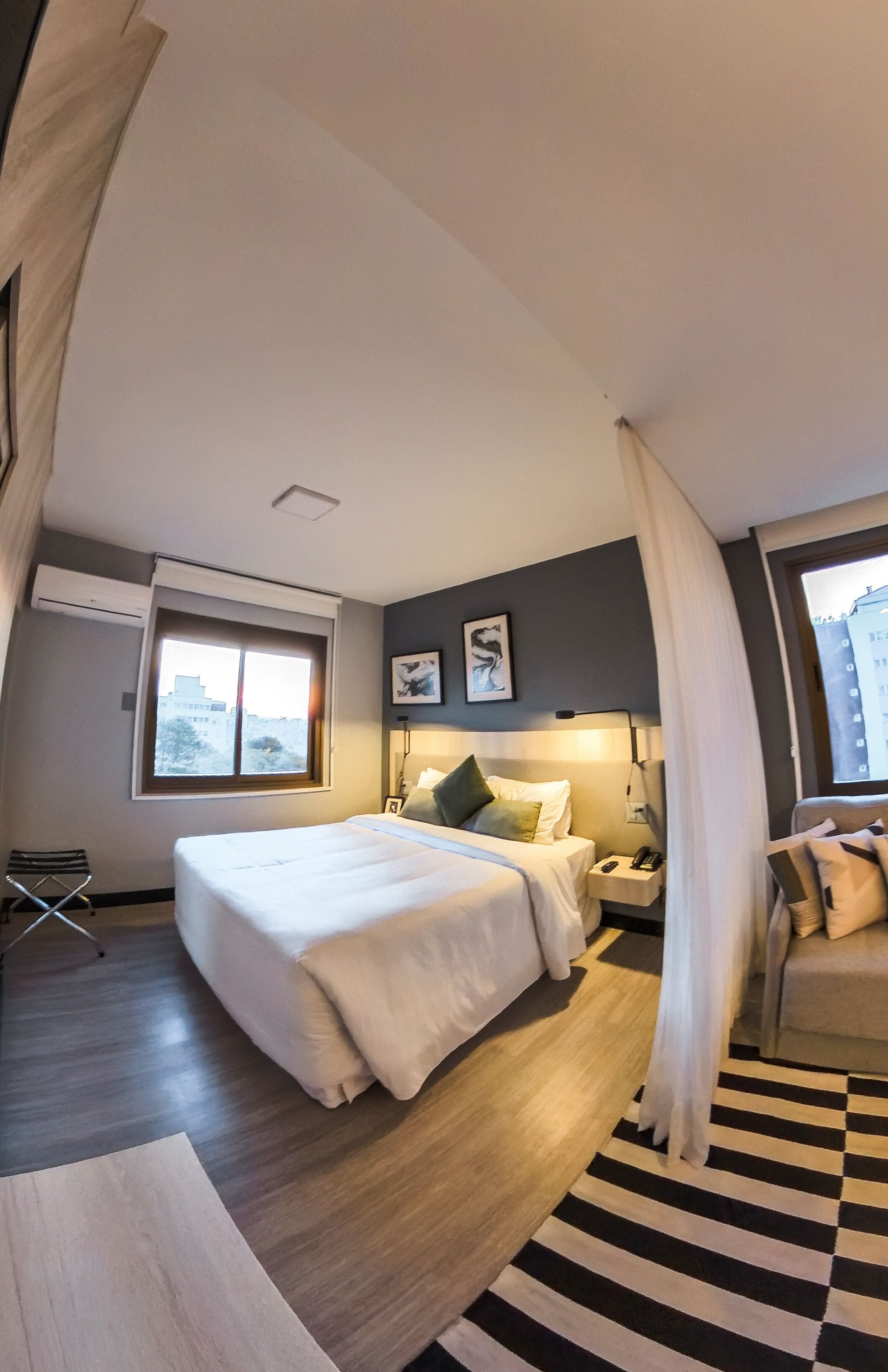 Une chambre d’hôtel avec un grand lit, une table de chevet, une chaise, une fenêtre avec des rideaux et une vue sur les bâtiments environnants.