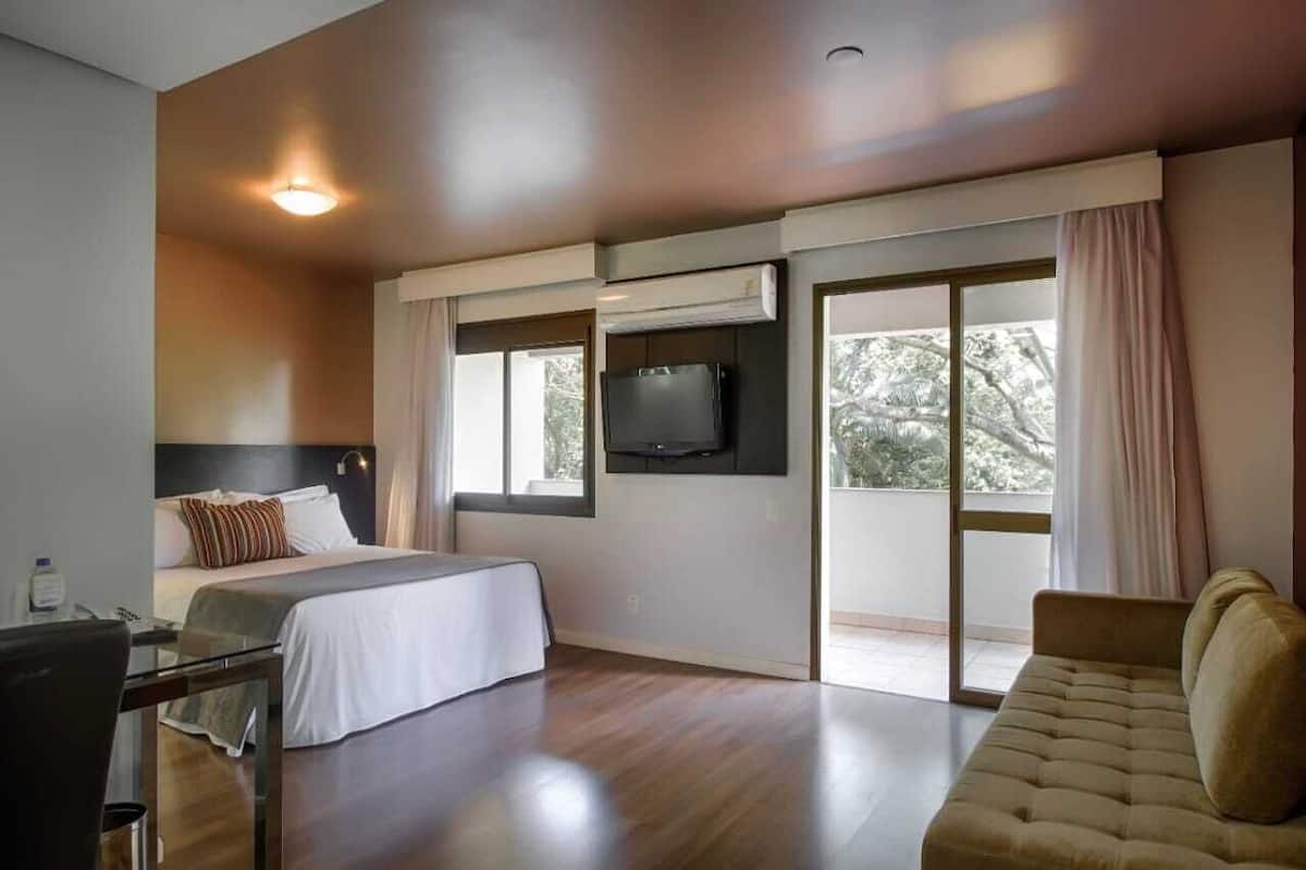 Una habitación de hotel moderna con una cama grande, un sofá, un televisor montado en la pared y una puerta corrediza de vidrio que da a un balcón.