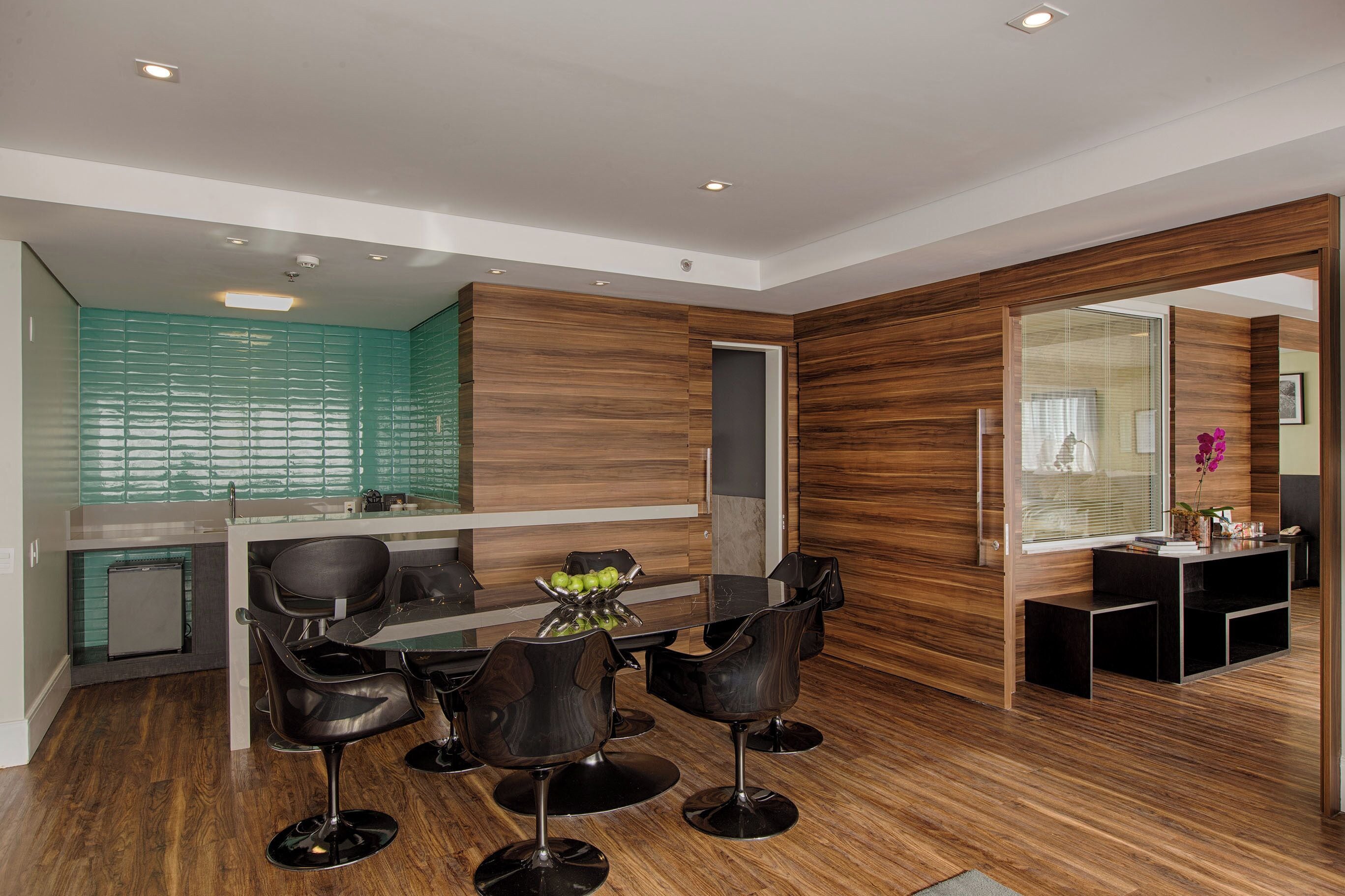Cozinha moderna com painel de fundo verde, painéis de madeira e área de jantar com mesa de vidro e cadeiras pretas.