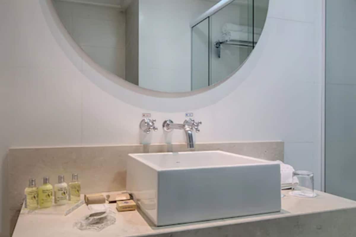Un baño moderno con un lavamanos blanco, un espejo redondo y una cabina de ducha de vidrio.