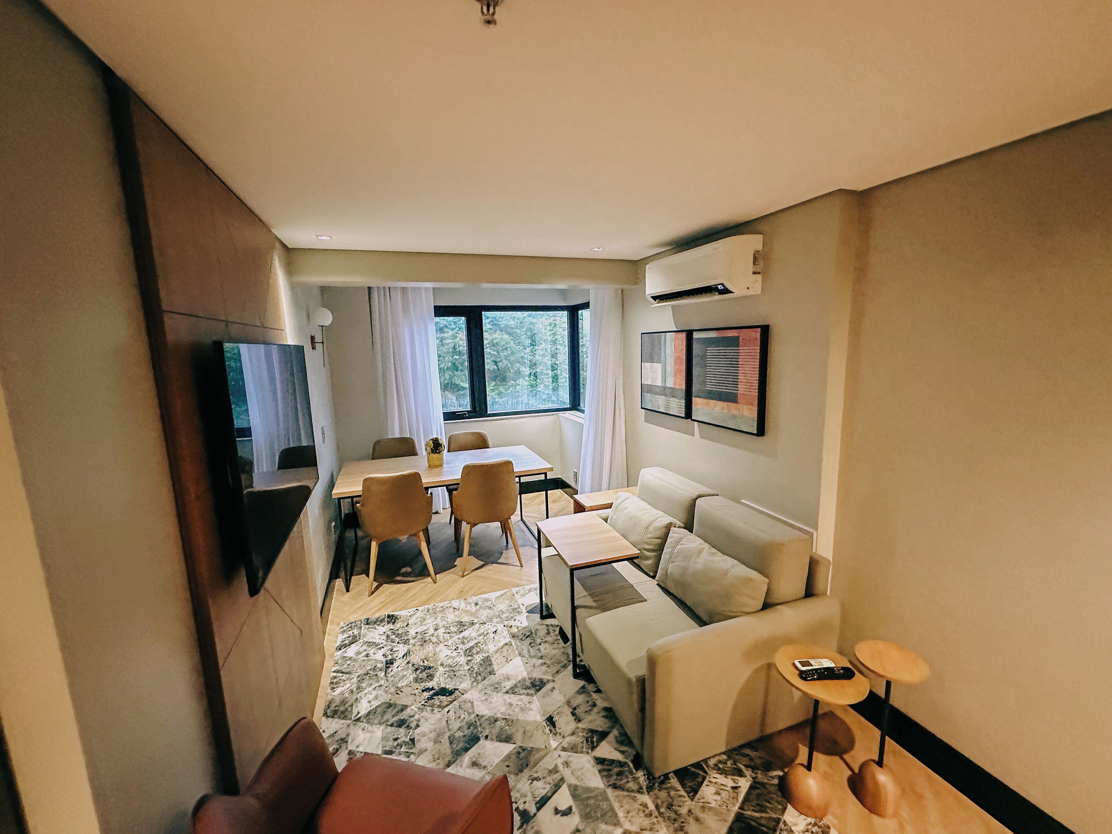 A modern hotel room with a dining area, a sofa, a TV, and a window with a view of trees.