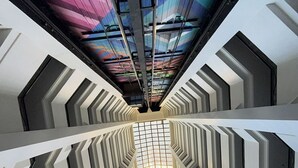 Um edifício moderno com um eixo de elevador de vidro central, paredes brancas geométricas e teto colorido.
