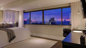 Quarto moderno com uma grande janela que oferece vista para a cidade ao entardecer.