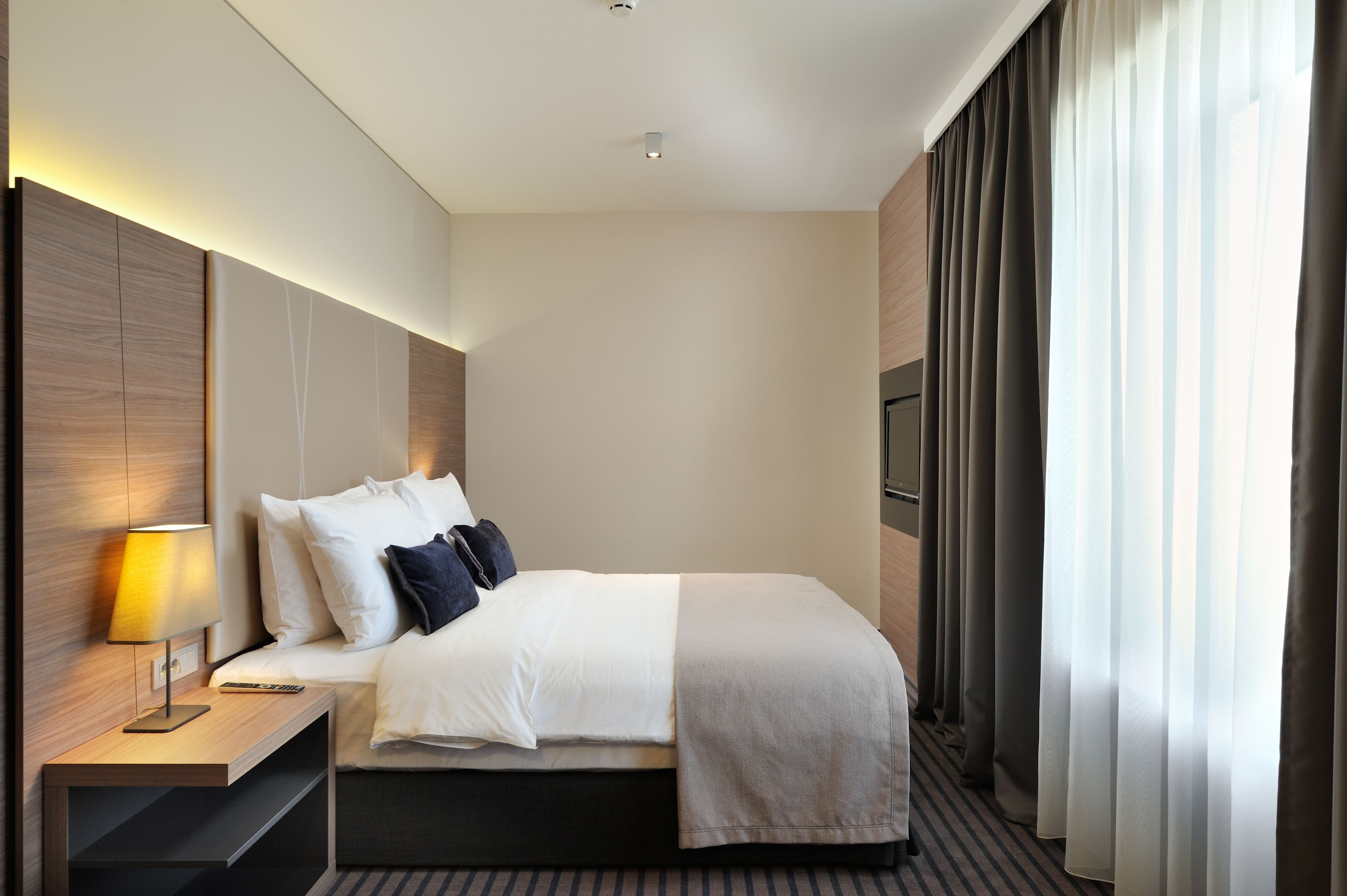 A modern hotel room with a neatly made bed, a bedside table with a lamp, and a large window with curtains.