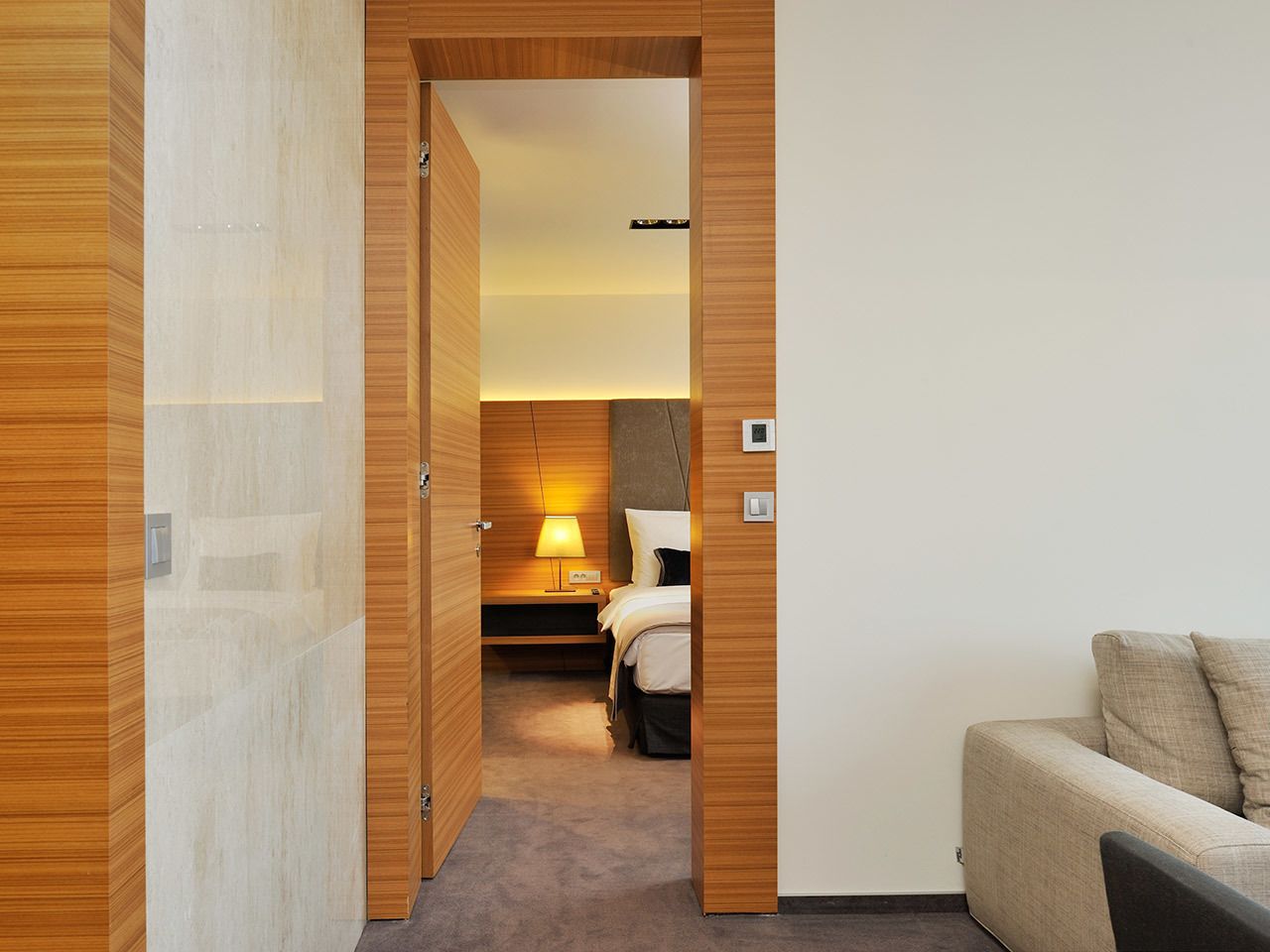 A hotel room with a wooden wardrobe, a bed with a nightstand, a lamp, and a sofa.