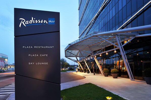 Ein modernes Gebäude mit einem Schild, das 'Radisson Blu' anzeigt, und Annehmlichkeiten wie einem Platzrestaurant, einem Café und einer Sky-Lounge.