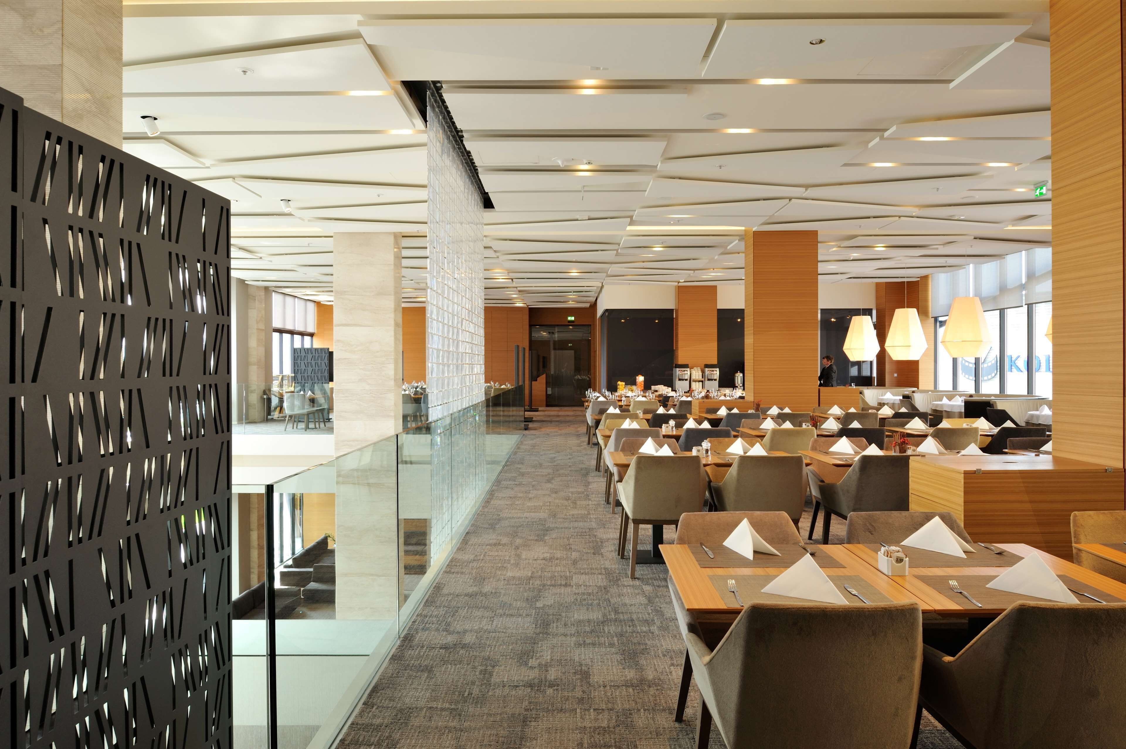 Un espace de restauration moderne dans un hôtel, doté de rampes en verre, de nombreuses tables et d’un éclairage contemporain.