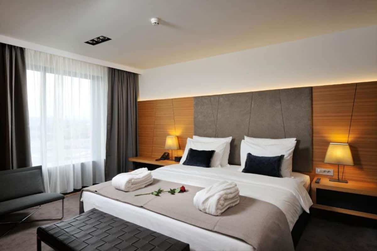 A hotel room with a large bed, two bedside lamps, a bedside table, a chair, and a window with curtains.