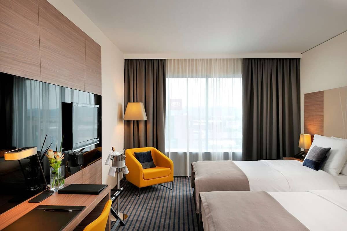 A hotel room with a bed, a desk with a laptop, a TV, a chair, and a window with curtains.