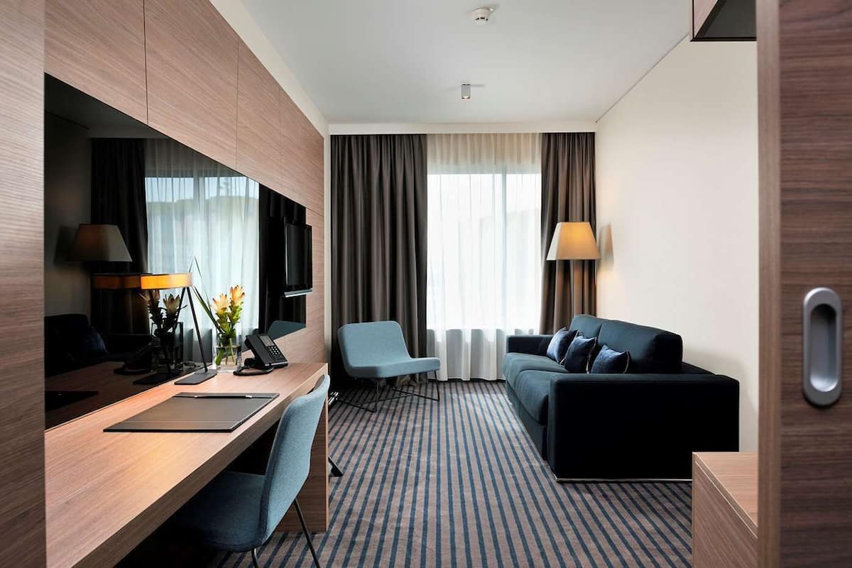 A modern hotel room with a wooden desk, a blue armchair, a black sofa, and a large window with curtains.