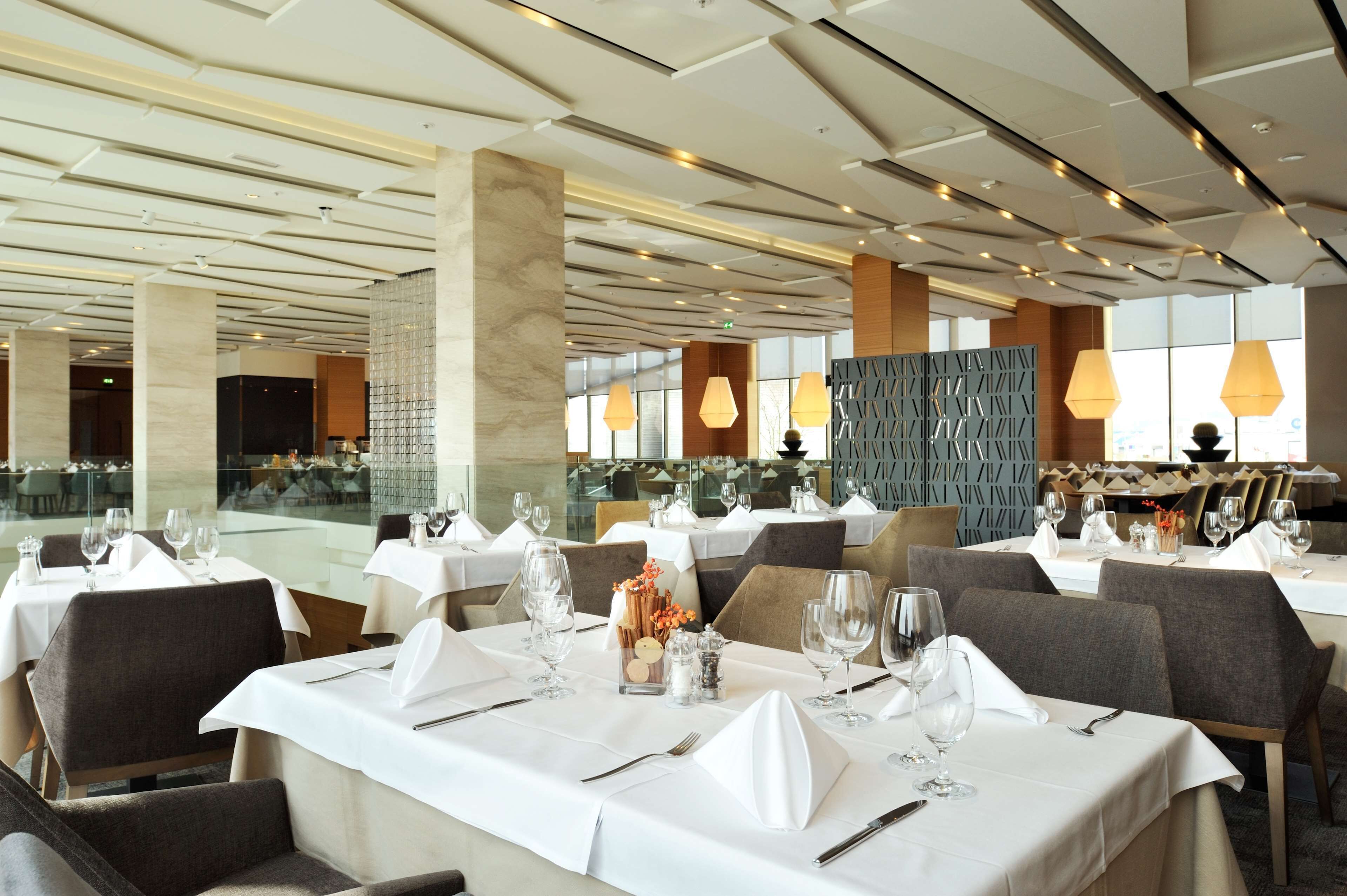 A modern dining area with tables set for service, large windows, and contemporary lighting fixtures.