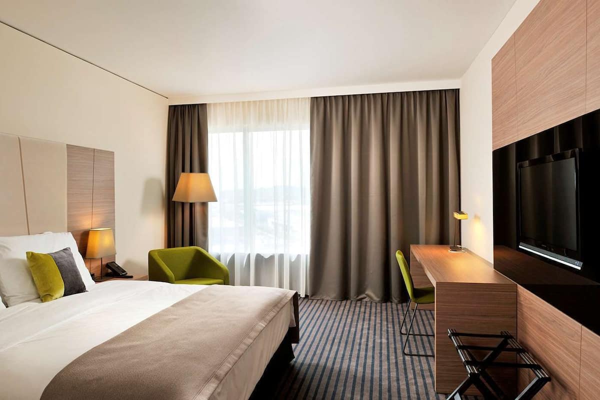 A hotel room with a large bed, a desk, a TV, and a view of the city through the window.