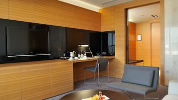 A modern hotel room with a wooden panel wall, a flat-screen TV, a desk with a laptop, a grey sofa, and a round coffee table with fruit.