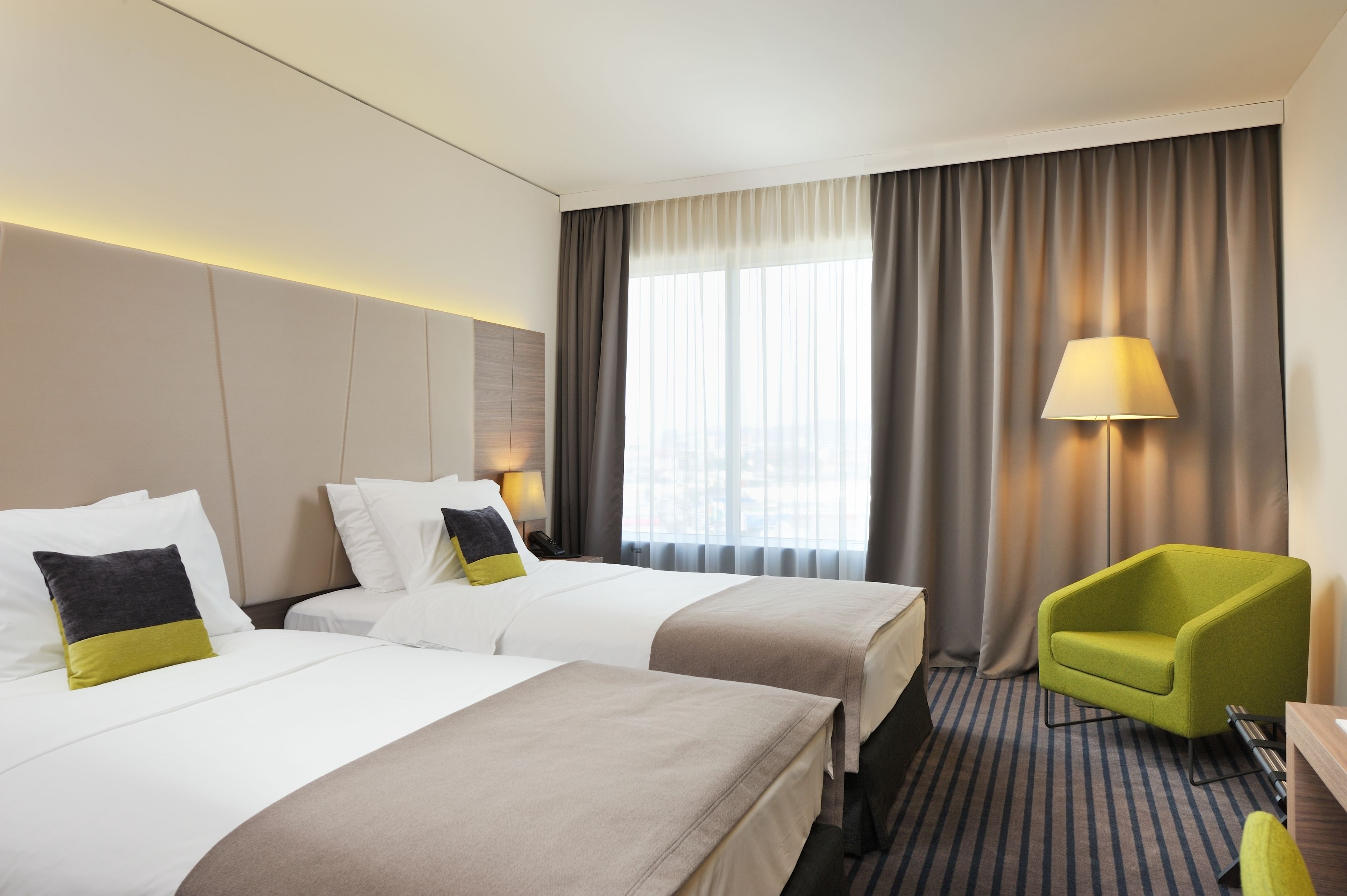 Ein Hotelzimmer mit zwei Betten, einem grünen Sessel, einer Lampe und Blick auf die Stadt durch große Fenster.