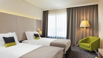 A hotel room with two beds, a green armchair, a lamp, and a view of the city through large windows.