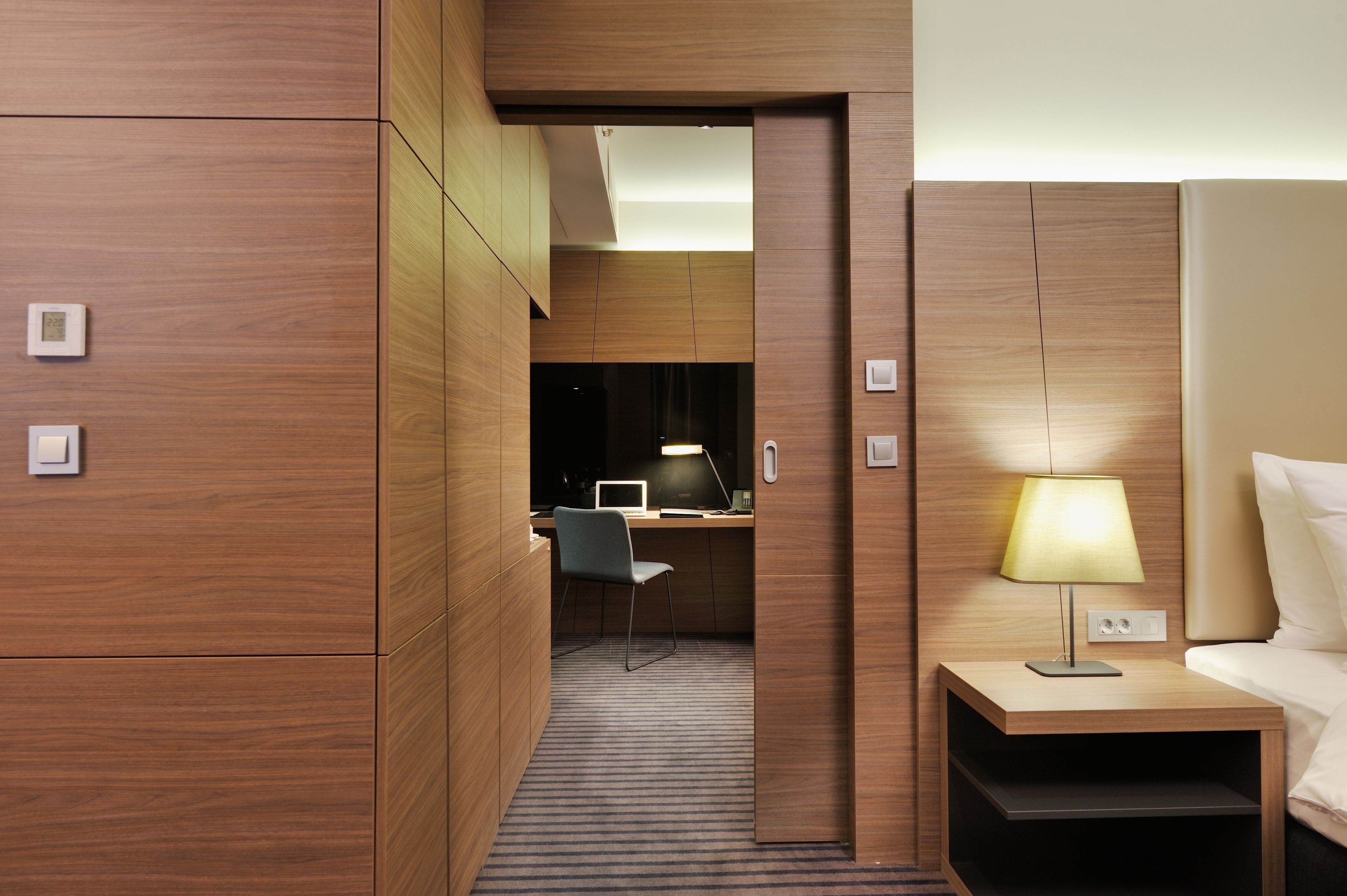 A hotel room with a wooden wall, a desk with a chair, a lamp, and a bed.