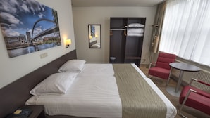 A hotel room with a bed, a red chair, a small table, a mirror, and a painting of a bridge.