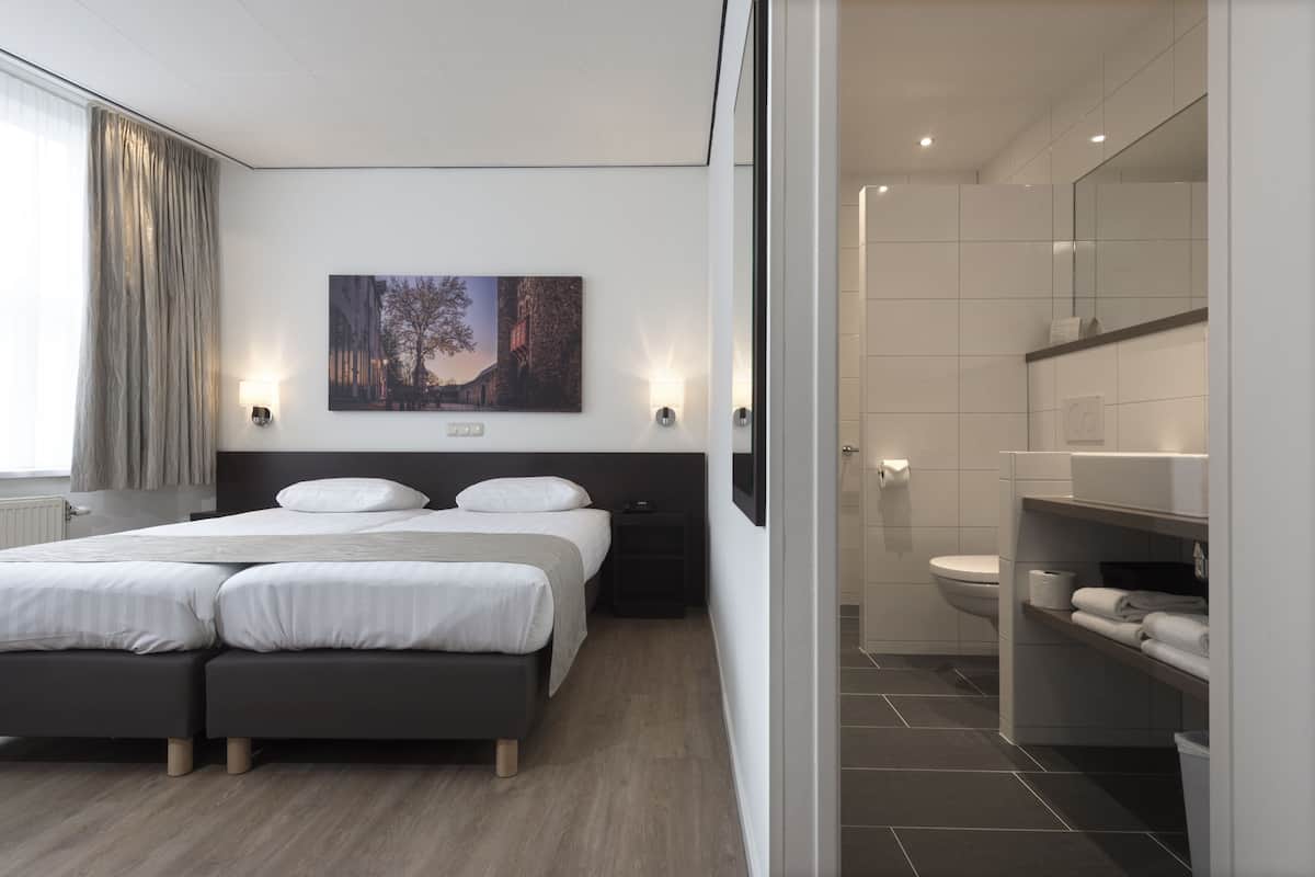 A modern hotel room with a large bed, a bedside table, a wall-mounted TV, and a bathroom with a toilet and shelves.