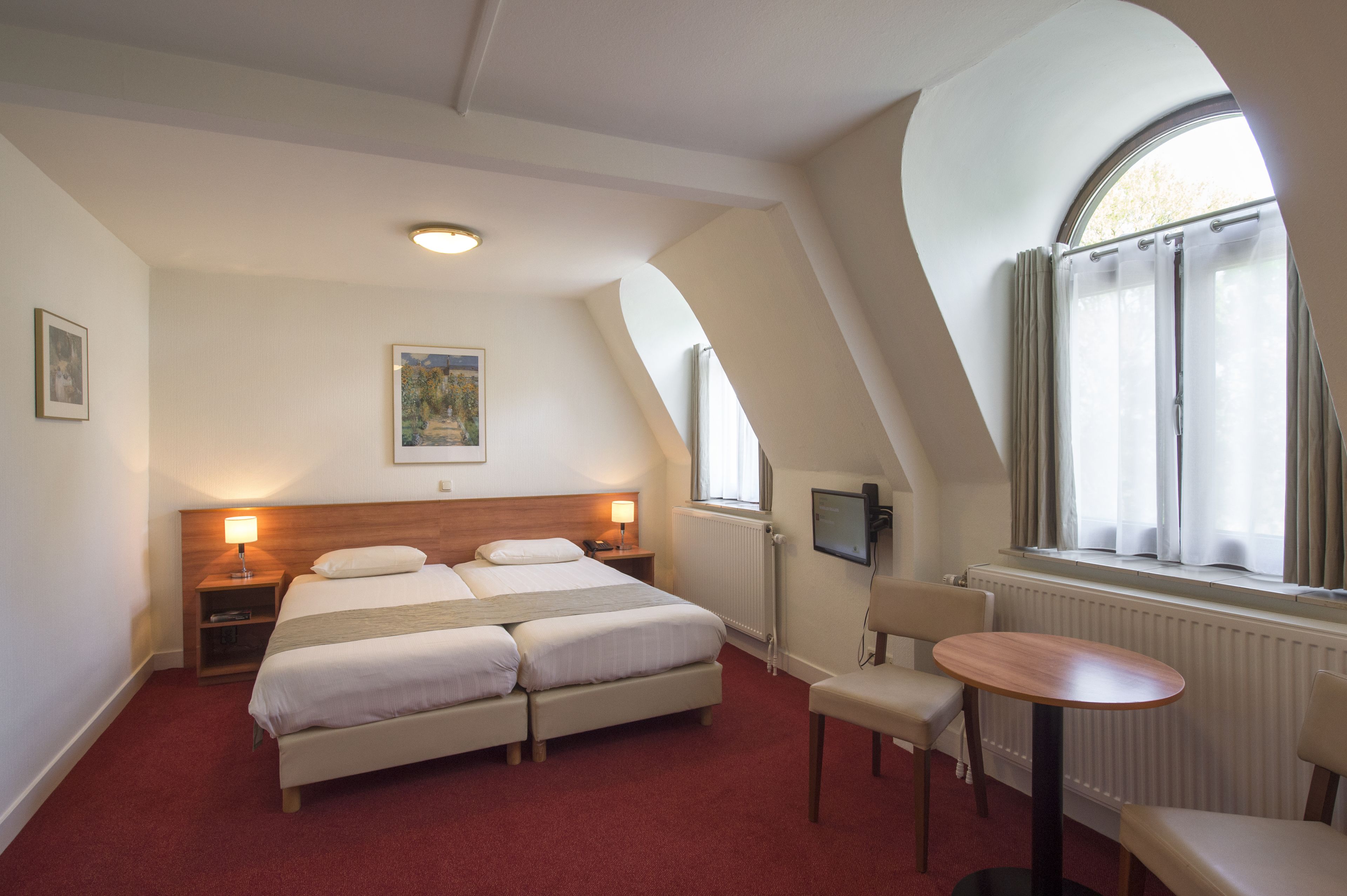 A hotel room with a double bed, two bedside tables with lamps, a small round table, a chair, a TV, and a window with curtains.