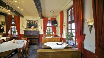 A dining area with wooden tables and chairs, a piano, and a television mounted on the wall.