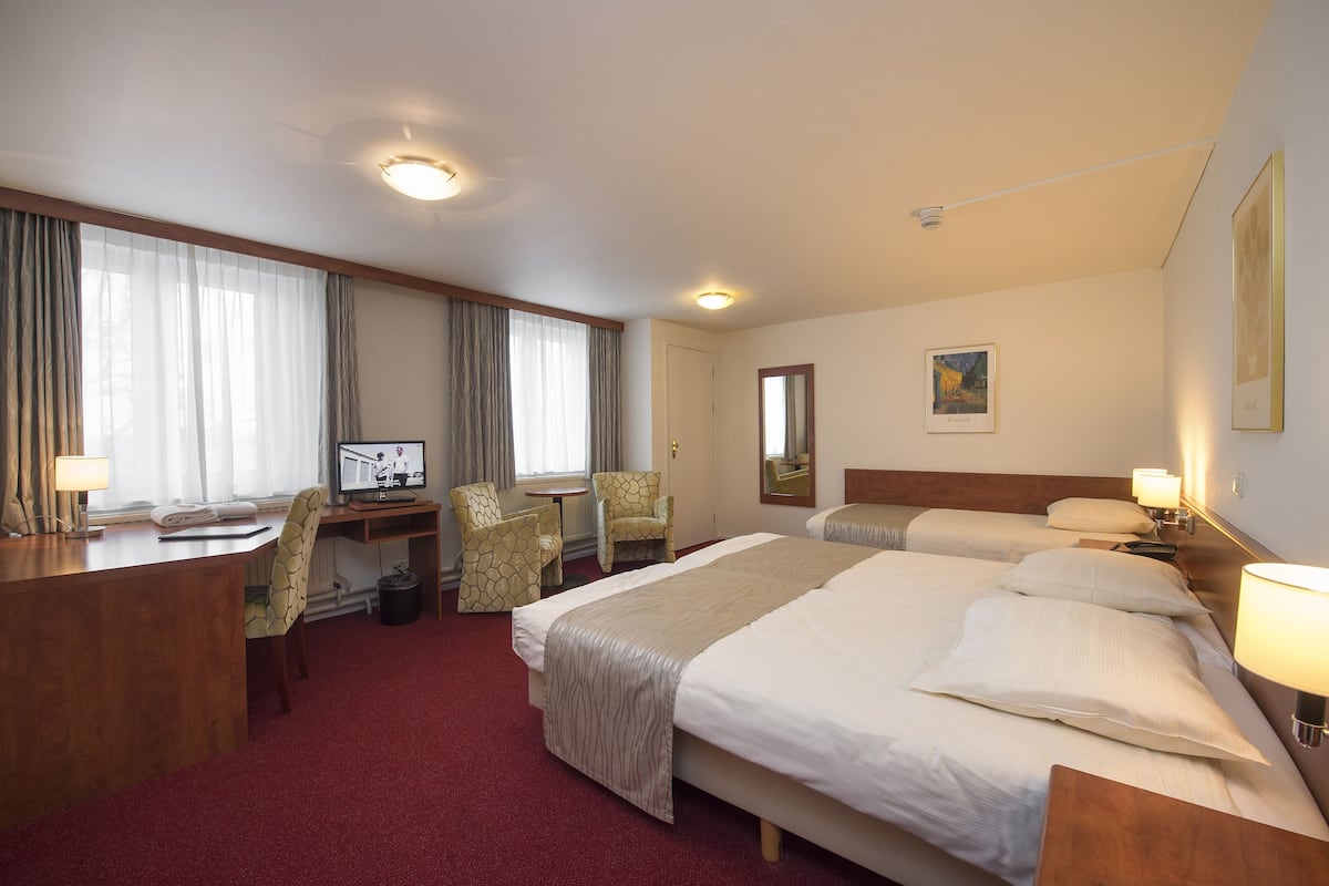 A hotel room with a large bed, a desk with a computer, two chairs, a small table, and a window with curtains.