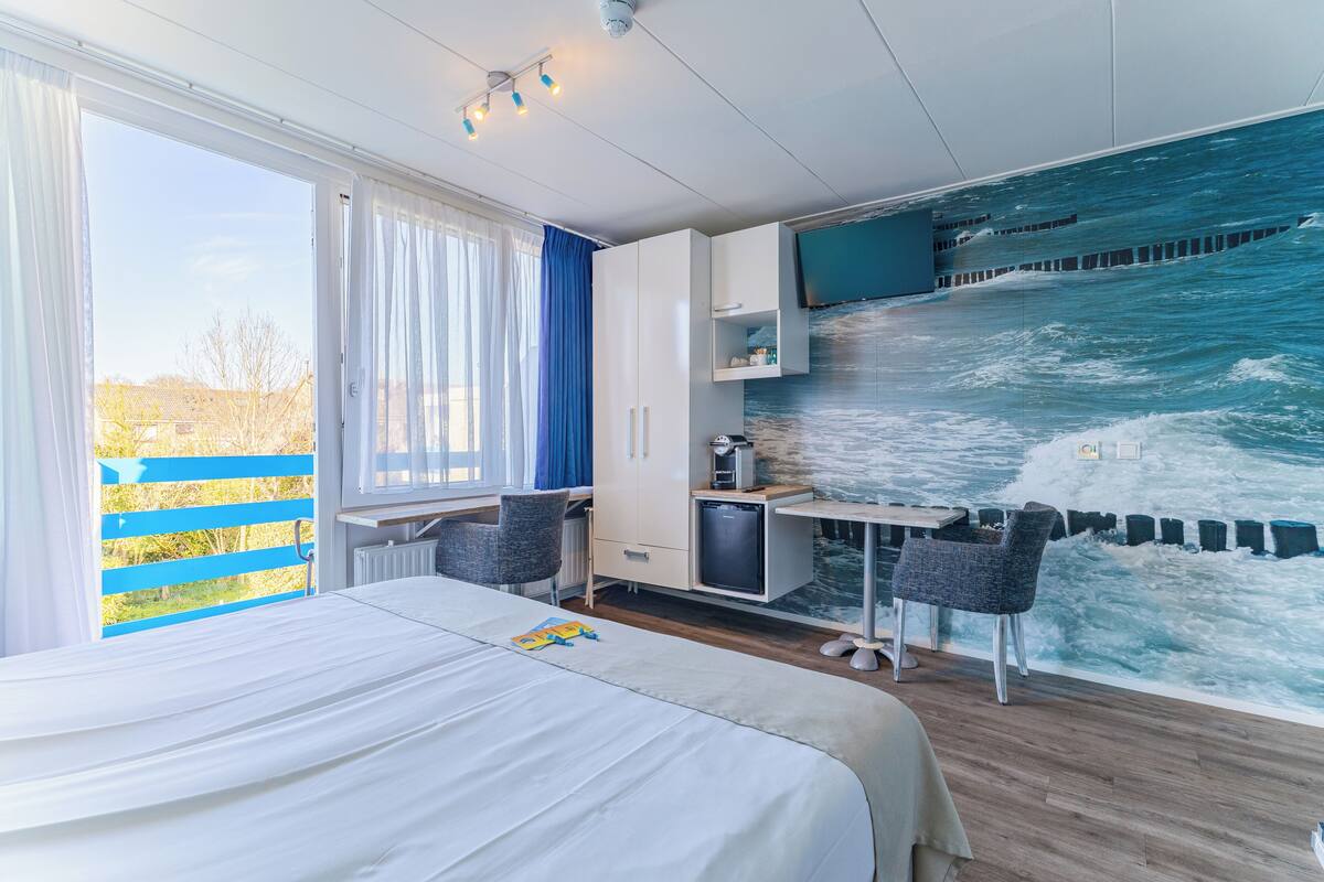 A bedroom with a bed, a desk, a chair, a wardrobe, and a large window with curtains.