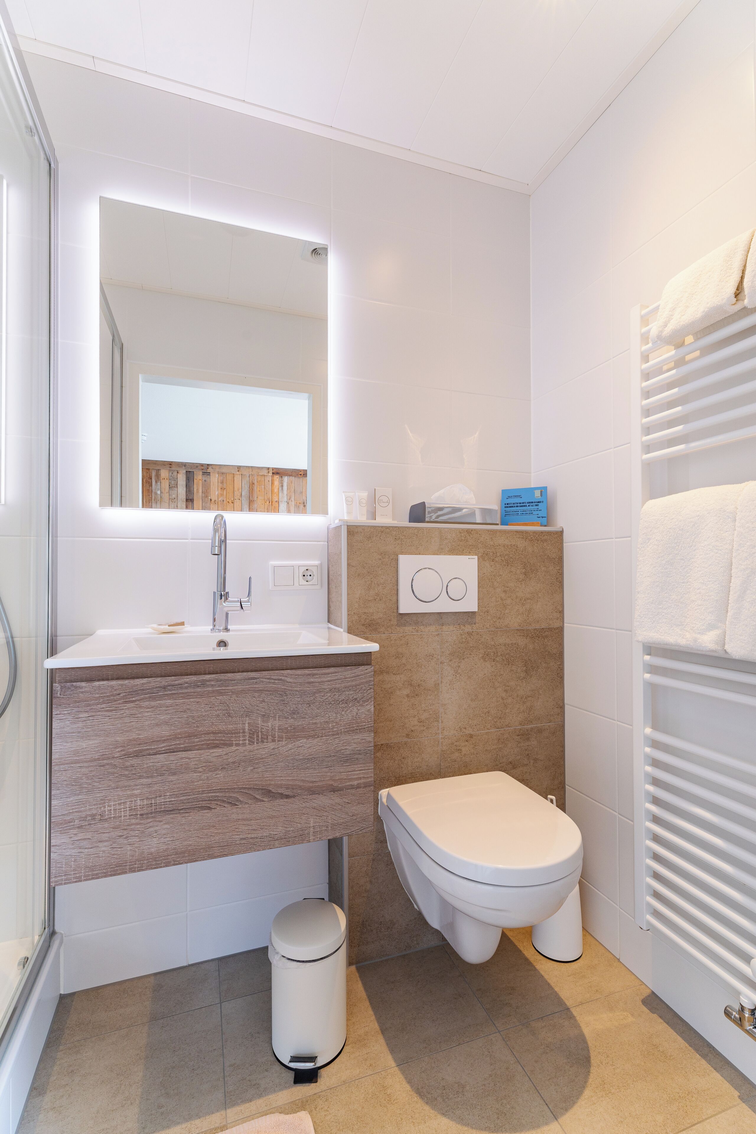 Un baño con lavamanos blanco, espejo, inodoro, toallero y un cubo de basura.