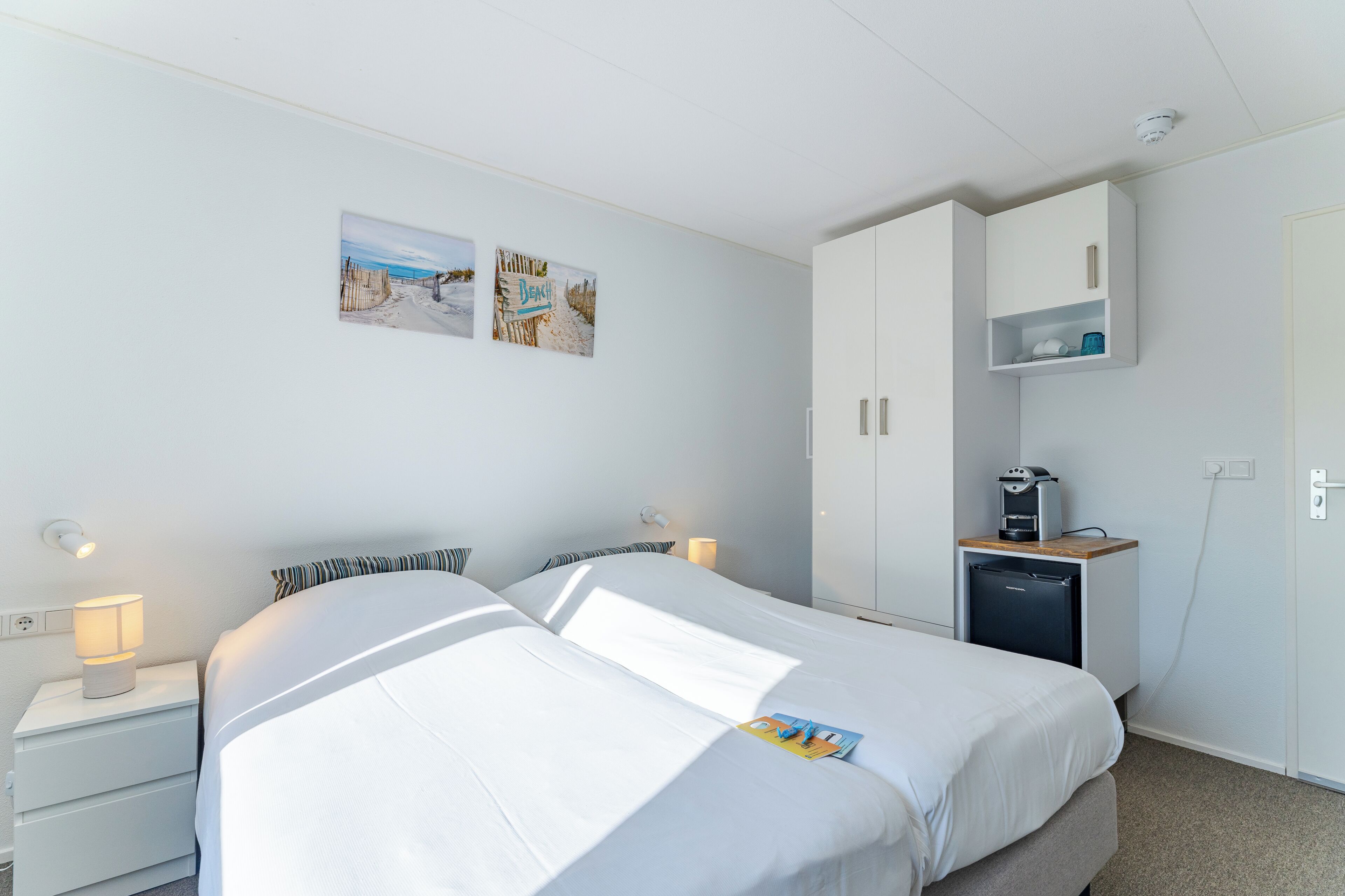 A bedroom with two beds, a nightstand with a lamp, a small refrigerator, and a cabinet with a shelf.